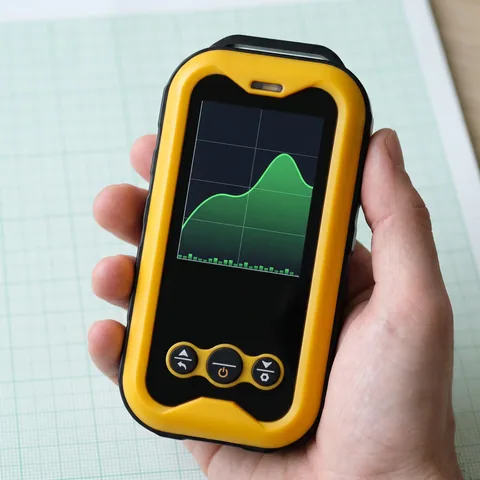 Close-up of the yellow Geiger counter's lit screen showing a green trend graph, held in one hand above graph paper